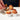 A hand decorates chocolate-frosted cupcakes from the American Dessert Masterclass, topping them with colorful edible flowers, presented in white paper cups on a white surface.